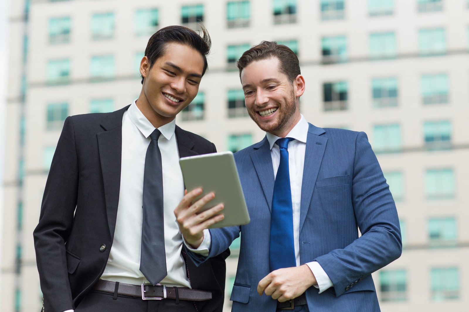 Closeup of two smiling young business men using tablet computer and standing with office building in background